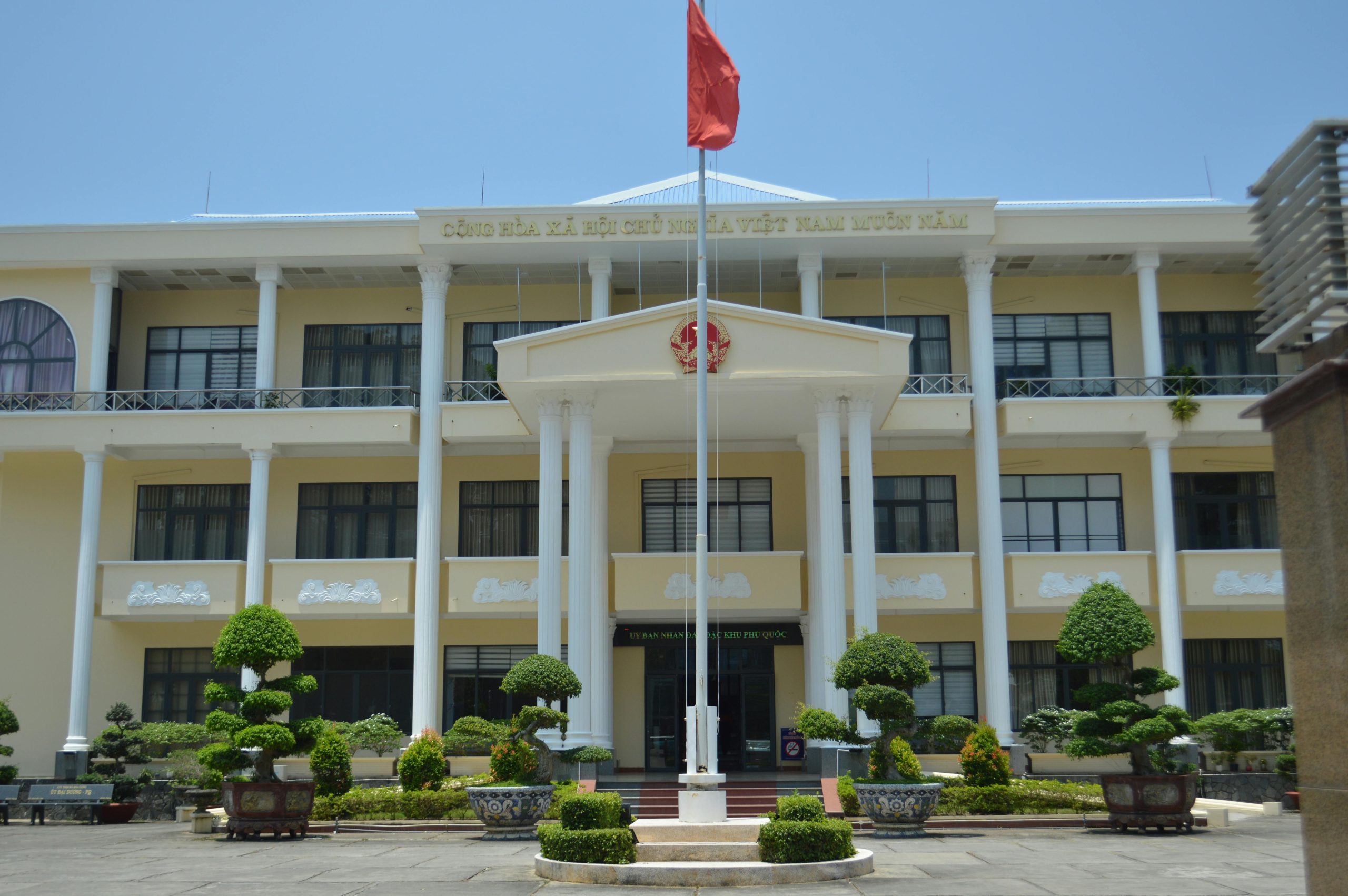 Front view of a government building in Phu Quoc, Vietnam, with a red flag and decorative plants.