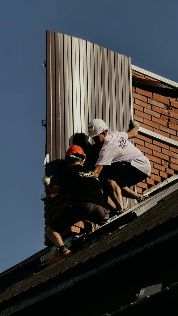 Two construction workers are installing metal panels on a brick building roof.